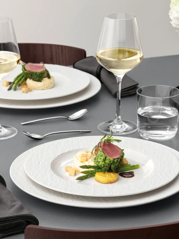 A formal table setting with a Manufacture plate on top of a plate A formal table setting with a white plate on a white plate with food on it, glassware, and silver cutlery on a grey table.