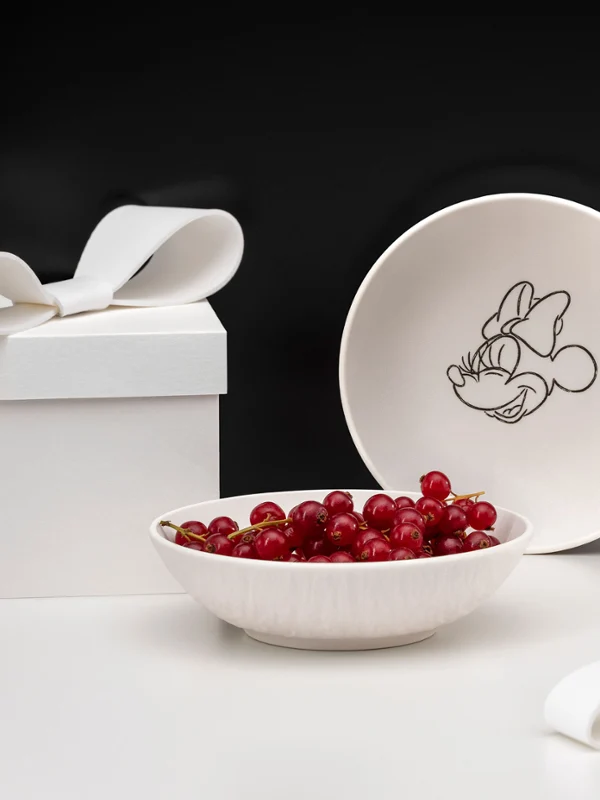 Dos cuencos blancos Manufacture Mickey con bayas rojas Dos bandejas blancas Manufacture Mickey, una con bayas rojas junto a una caja de regalo blanca con un lazo sobre una superficie blanca.