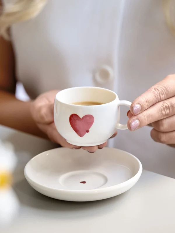 Filiżanka do kawy i spodek marki with love z motywem czerwonego serca Osoba trzyma białą filiżankę do kawy z motywem czerwonego serca nad dopasowanym spodkiem z motywem czerwonego serca na stole.