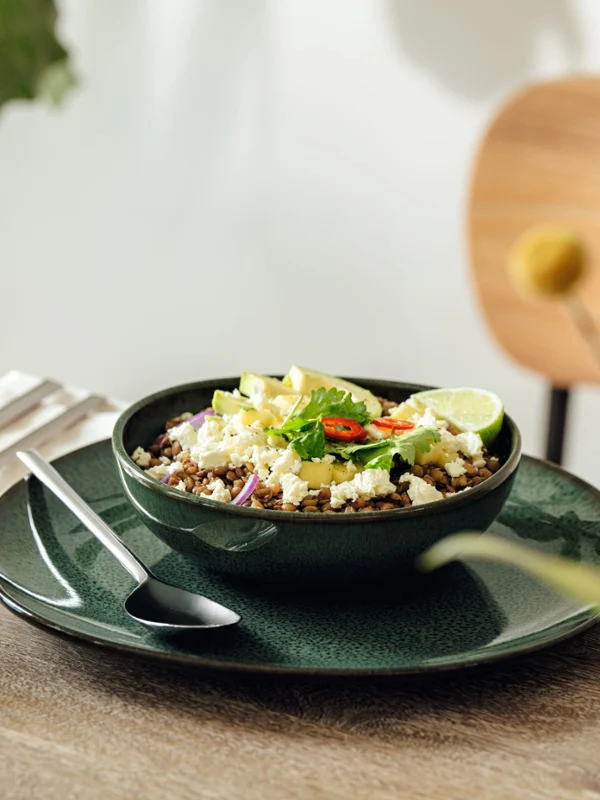 A bowl of salad with avocado, lime and chili is served in a dish on a wooden table set for a meal.