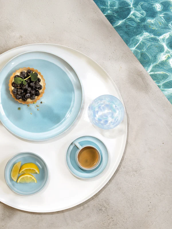 A round tray with tableware from the Perlemor Aqua collection and a glass of water, at the edge of a pool.