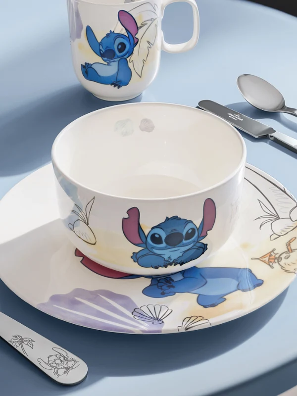 Una mesa puesta con la colección Disney Stitch sobre un fondo azul. Un cuenco, un plato, una taza, un tenedor, una cuchara y un cuchillo con motivos de Stitch sobre una mesa azul bajo luz natural.