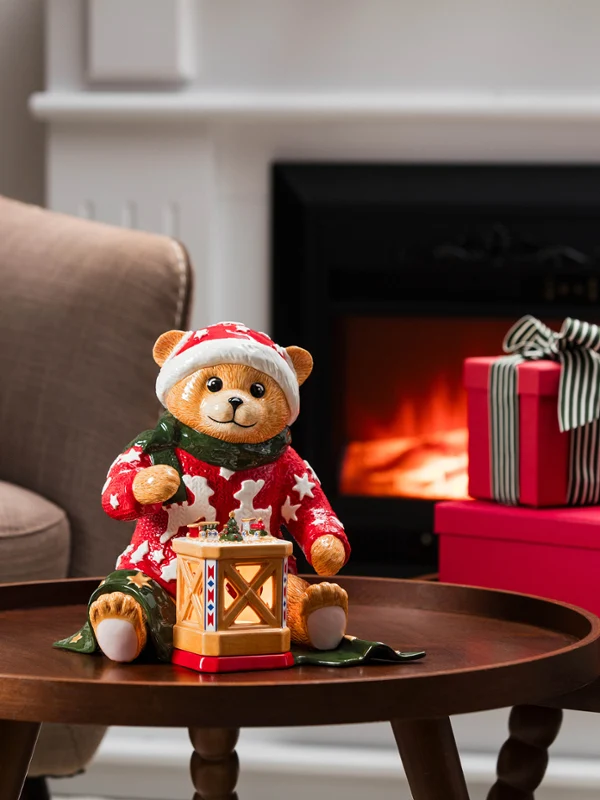 A teddy bear in pajamas, holding a lantern from Villeroy & Boch, sits on a coffee table next to gifts and flowers in a living room with a lit fireplace.