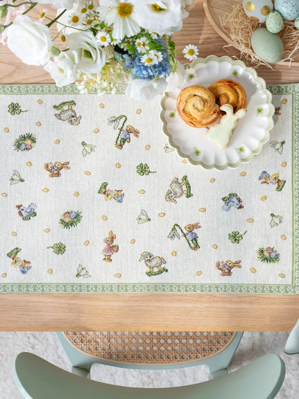 A wooden table set with a Easter Delight placemat, pastries on a plate, flowers, dyed eggs, and a folded napkin.