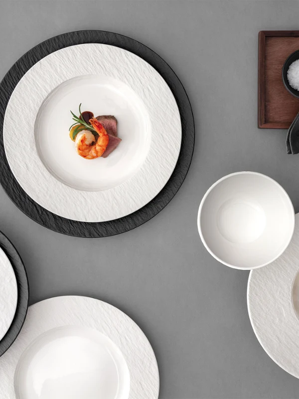 A formal table setting with a white and black plates, with food on it on a grey table.