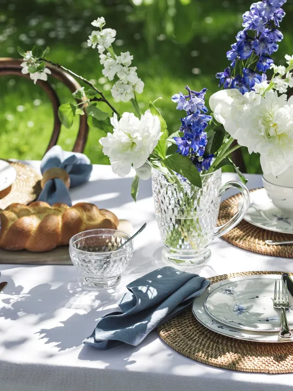 A garden table setting with Villeroy & Boch "Old Luxemburg" floral porcelain, wicker placemats, blue-tinted glass tableware, a crystal jug with blue and white flowers and a loaf of bread.