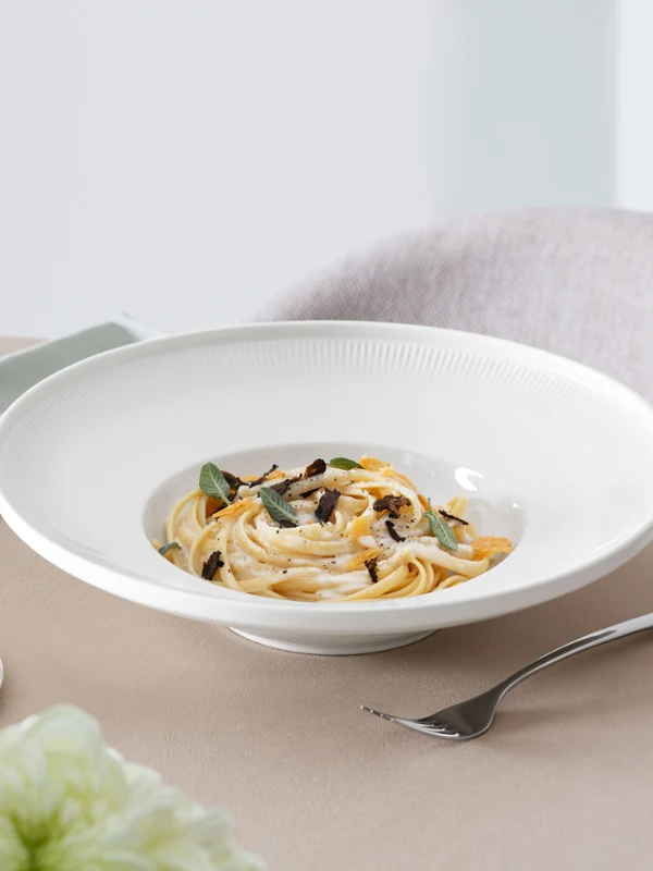 Une assiette à pâtes Afina de Villeroy & Boch avec des tagliatelles, de la sauge et de la sauce blanche, à côté d’un verre à vin blanc ainsi que de couverts de table Piemont.