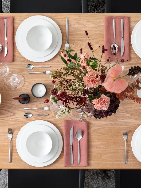 Un tavolo da pranzo in legno con piatti bianchi, ciotole, posate su tovaglioli rosa, due candele, una ciotola con uva e una composizione floreale.