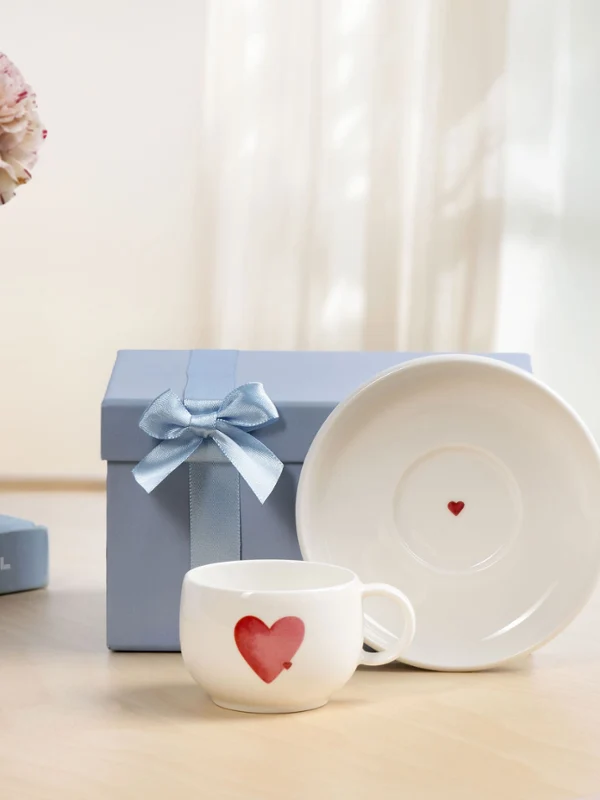 Filiżanka z podstawką With Love jako pomysł na prezent Biała filiżanka With Love z czerwonym motywem serca, dopasowana podstawka i niebieskie pudełko prezentowe z kokardką na drewnianym stole przy oknie.