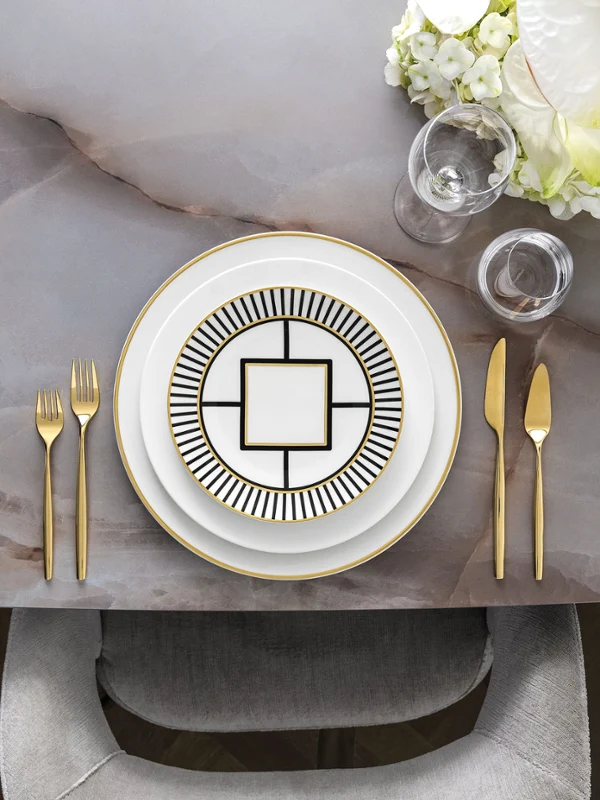 Stilvolles Essgeschirr mit elegantem MetroChic Geschirr, goldenem Besteck und einem floralen Tafelaufsatz auf einem Marmortisch.