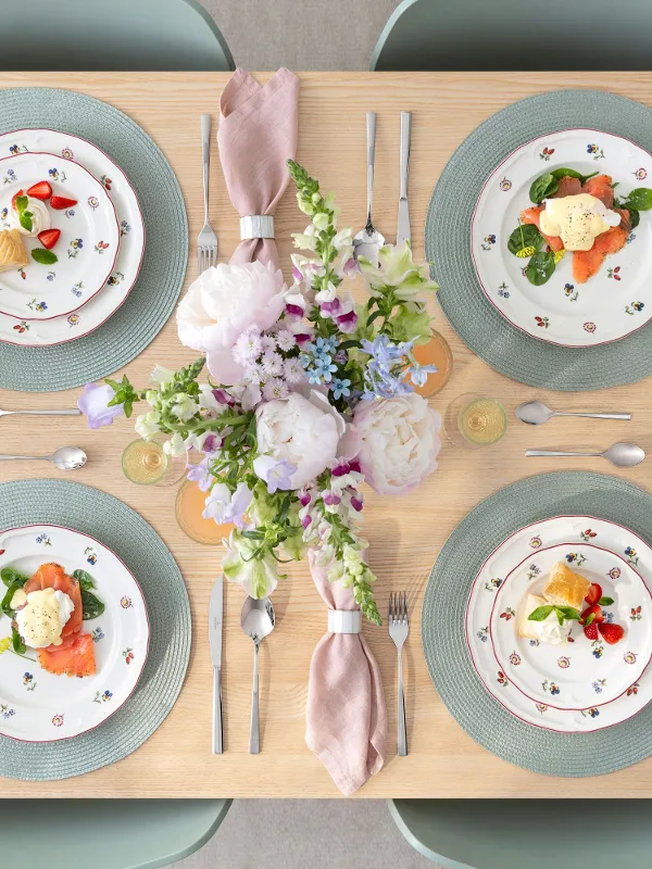 A set dining table for four people with Petite Fleur flower plates, pink napkins, drinks, and a floral arrangement.