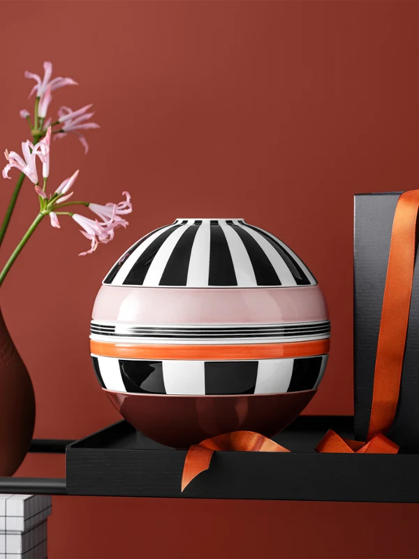 A striped decorative La Boule Memphis A striped decorative La Boule Memphis, a black box with an orange ribbon, and a brown vase with pink flowers are arranged on a shelf against a red background.