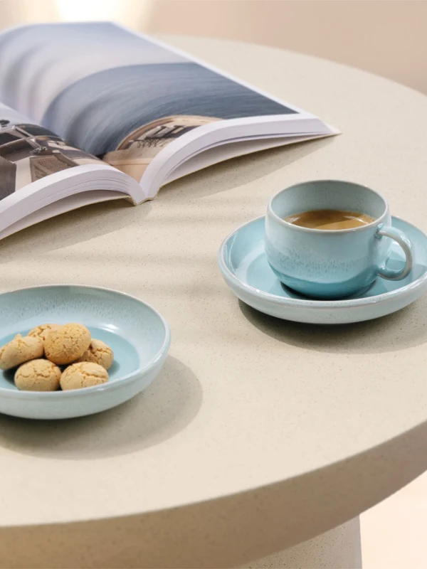 A Perlemor Aqua coffee cup, a plate of cookies on a table A Perlemor Aqua coffee cup, a plate of cookies, and an open magazine are on a round, light-colored table.
