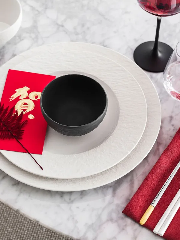 Une table en marbre blanc avec des assiettes Manufacture blanches, un plat creux Manufacture noir, une enveloppe rouge et un décor de fougère rouge. Un décor de style asiatique.