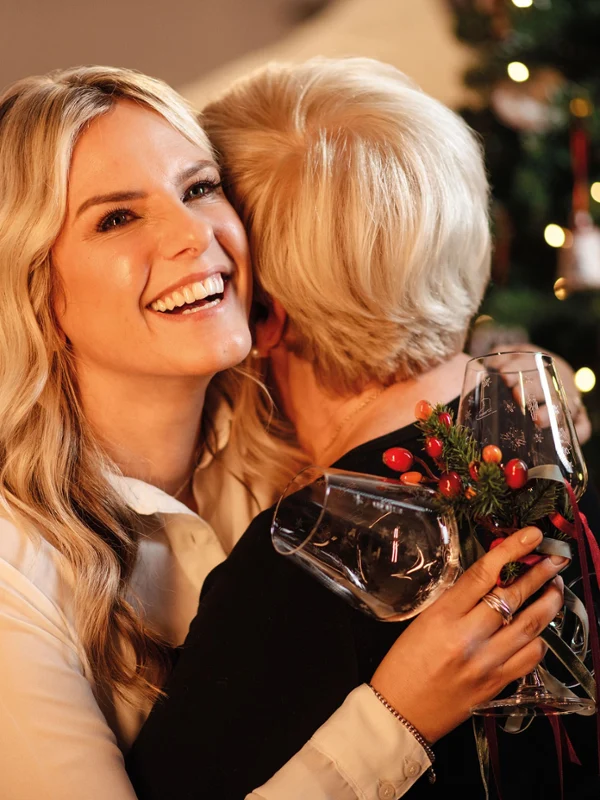 Two women hugging, one holding a Toy's Delight wine glass. Two women hugging in front of a decorated Christmas tree. The woman looking into the camera is holding a Toy's Delight wine glass.