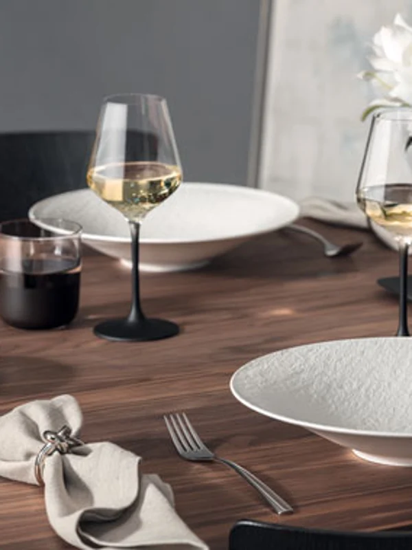 Table à manger Manufacture avec assiettes, verres et vase Une table à manger composée d'assiettes blanches Manufacture, de verres à vin, de couverts et d'une serviette. Un vase de fleurs blanches Manufacture se trouve à l'arrière-plan.