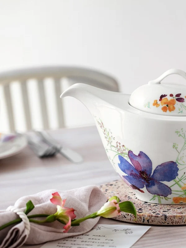 Kwiecisty czajniczek Villeroy & Boch stoi na korkowej macie Czajniczek w kwiaty od Villeroy & Boch stoi na korkowej macie, otoczony pasującymi filiżankami na stole jadalnym z białymi krzesłami w tle.