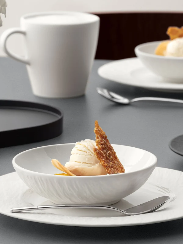 A formal table setting with a white bowl on a white plate with food on it, cup, and silver cutlery on a grey table.