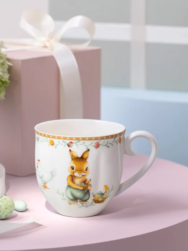 A decorated Annual easter Edition mug sits on a pink surface A decorated Annual easter Edition mug sits on a pink surface next to a white ribbon, pastel gift boxes, and a vase of flowers.