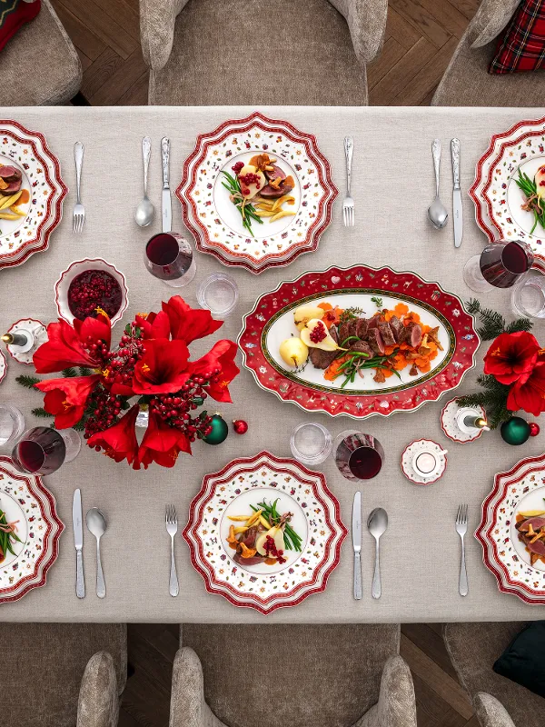A festive dining table setting for eight people with red and white patterned plates, red flowers, glasses, cutlery and various dishes, including pasta and salad, on a white tablecloth.