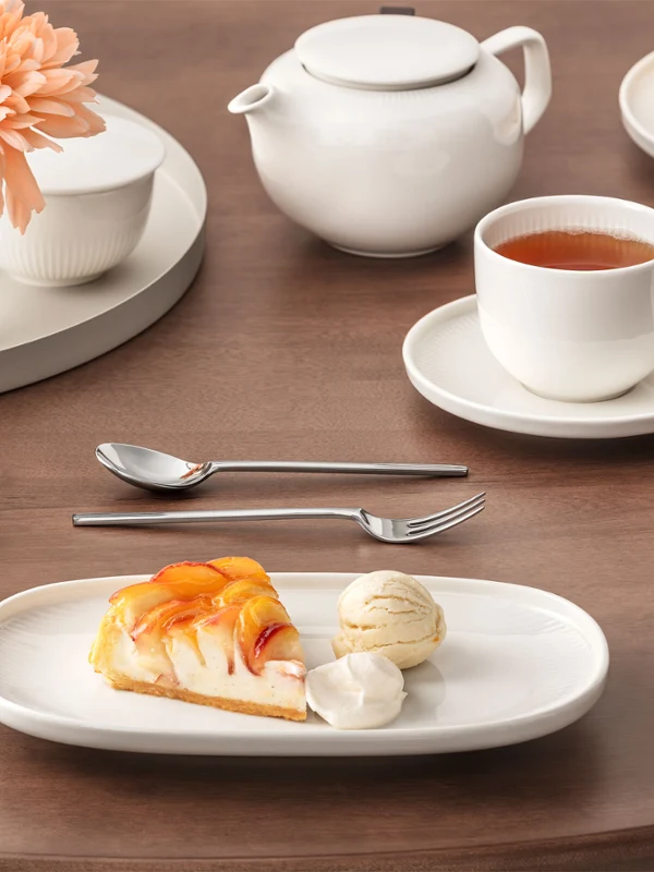 Der gedeckte Tisch der Afina Kollektion zeigt eienn Teller mit Kuchen und Eiscreme, Tasse Tee und eine Teekanne stehen auf einem Holztisch mit einer Blumenvase.
