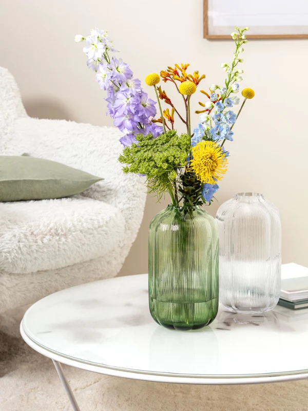 A Villeroy & Boch Fleur vase with colourful flowers and a green vase on a round marble table A round coffee table with a Villeroy & Boch Fleur vase holding blue and green flowers. In the background, an open magazine lies on a white, fluffy chair.