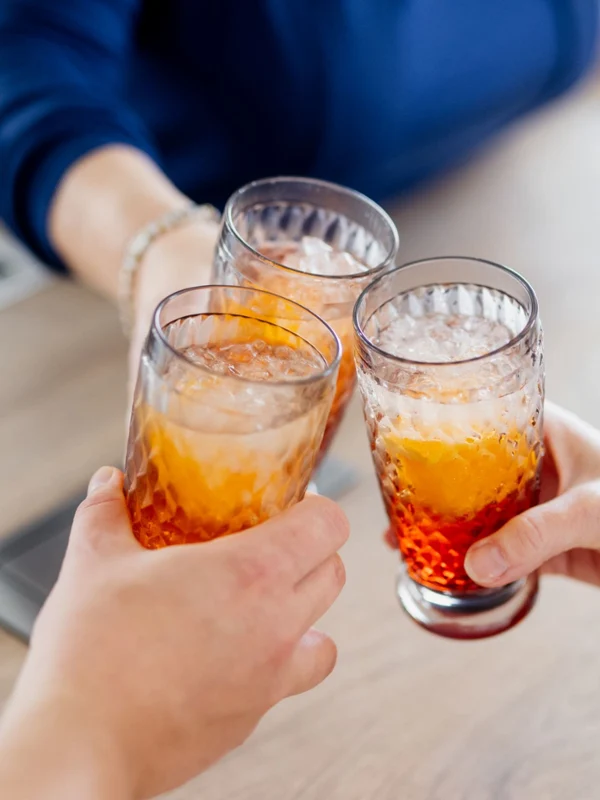 Szklanki Highball Boston Trzy osoby wznoszą toast szklankami wypełnionymi napojem pomarańczowym nad drewnianym stołem.