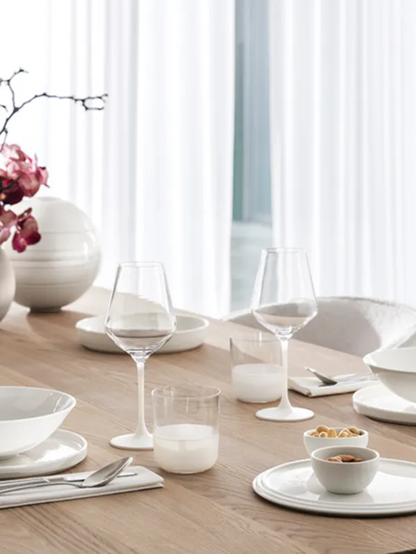 Un tavolo da pranzo moderno con stoviglie bianche, bicchieri e un vaso di fiori rosa su una superficie di legno.