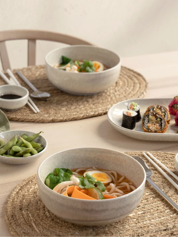 A table set for two with Perlemor pieces A table set for two with bowls of ramen, sushi rolls, edamame, chopsticks and dipping sauces from the Perlemor collection on woven placemats.