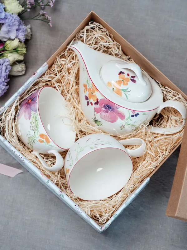 A Mariefleur tea set in a straw-filled gift box filled, next to an open invitation card and purple flowers.
