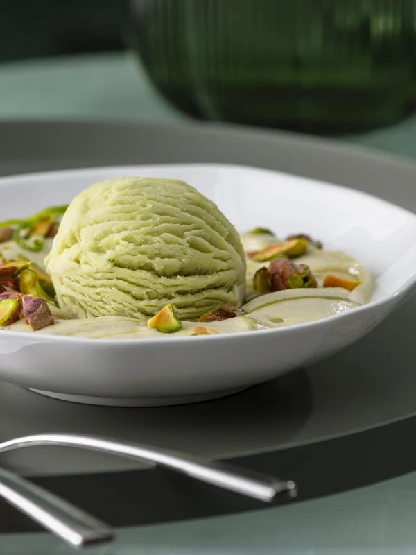 Eine Pura Dessertschale mit Pistazieneis Eine Pura Dessertschale mit Pistazieneis, gehackten Pistazien und Sahne auf einem weißen Teller mit Besteck daneben.