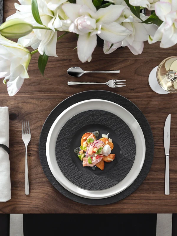 Manufacture Rock & NewMoon – Modern Contrast, Pure Character Vue de dessus d’une table contemporaine combinant la collection Manufacture Rock en noir mat—avec une assiette gourmet, un plat rectangulaire et une assiette à déjeuner—et la ligne minimaliste NewMoon en blanc éclatant, comprenant une assiette plate, une assiette à pain et un bol à sauce. Les formes et surfaces contrastées créent une composition artisanale harmonieuse et frappante.