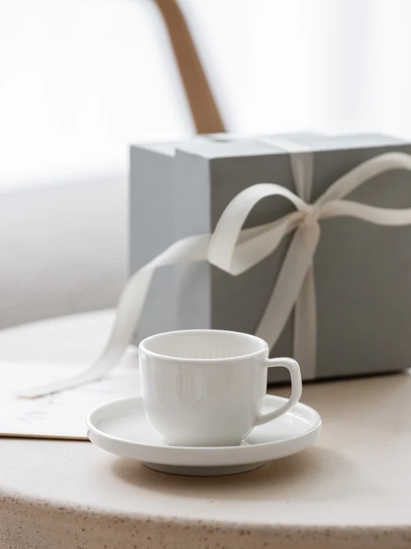 Een wit Afina kopje op een schoteltje, een cadeaubox met een strik en een boeket witte bloemen op een ronde tafel.