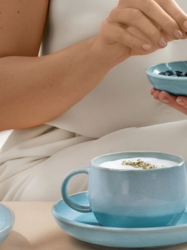 Perlemor Aqua bowl and coffee cup Person holding blueberries above a blue mother-of-pearl aqua bowl, with a blueberry pie and a Perlemor Aqua cup of coffee on a table in front of them.