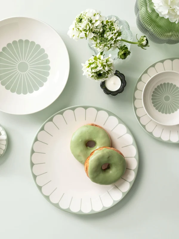 Plate with a green and white pattern on a light green surface with two green donuts and a small bouquet of white flowers.	