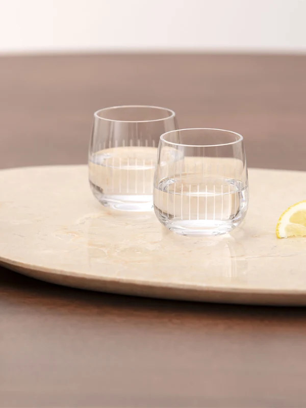 Zwei klare Wassergläser der Afina Kollektion, zwei Zitronenspalten und eine Schale mit Nüssen stehen auf einem runden Tablett auf einem Holztisch. Im Hintergrund ist eine Vase mit einer pfirsichfarbenen Blume zu sehen.