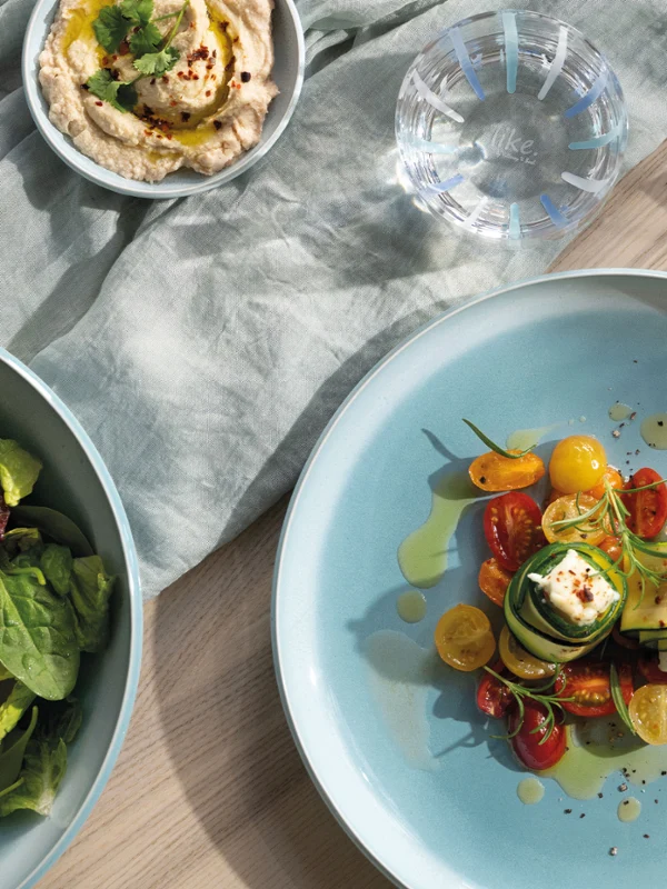 A Perlemor Aqua plate on a laid table A mother-of-pearl aqua plate with various tomatoes and cheese, a bowl of salad, hummus, olives, cutlery and a napkin on a table.