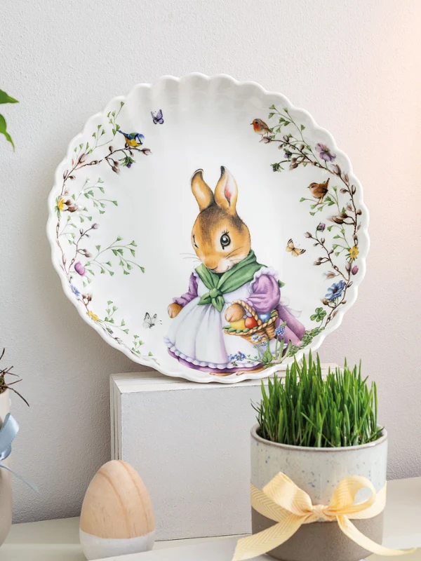 The Spring Fantasy decorative plate by Villeroy & Boch with a bunny motif is displayed on a shelf, surrounded by flowers, candles, and a wooden egg.