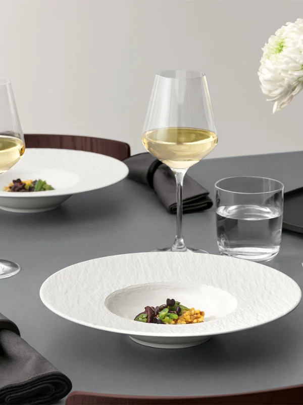 A formal table setting with a Manufacture pasta plate A formal table setting with a pasta plate ad food in it, glassware, and silver cutlery on a grey table.