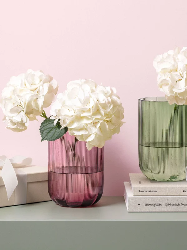 Green and pink glass vases by Fleur on a shelf Two colored glass vases by Fleur with white hydrangeas on a shelf, next to a gift box, books, and a small green lamp.