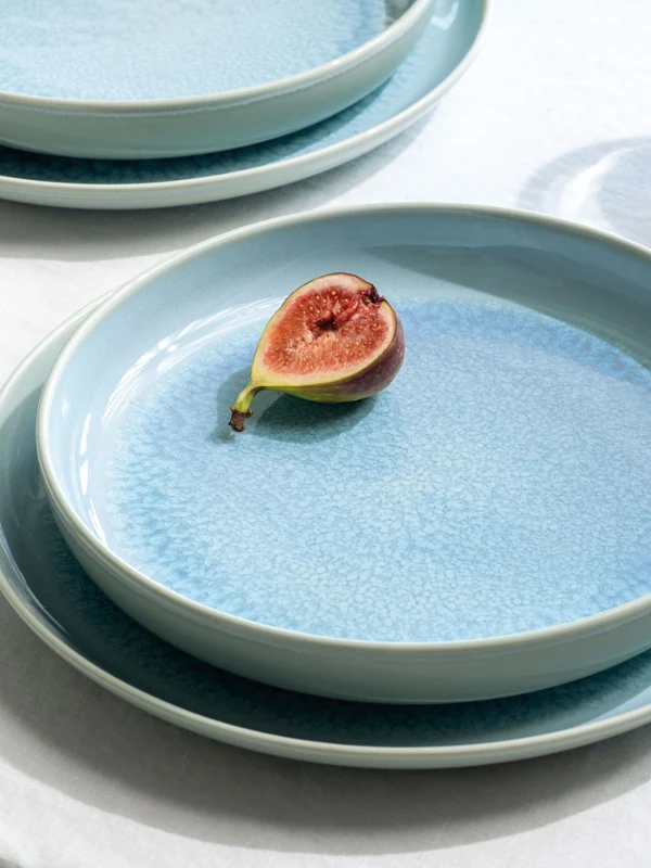 Eine Feige auf einem Crafted Teller auf einem weißen Tisch neben einem Glas Wasser und einer gestreiften Serviette.