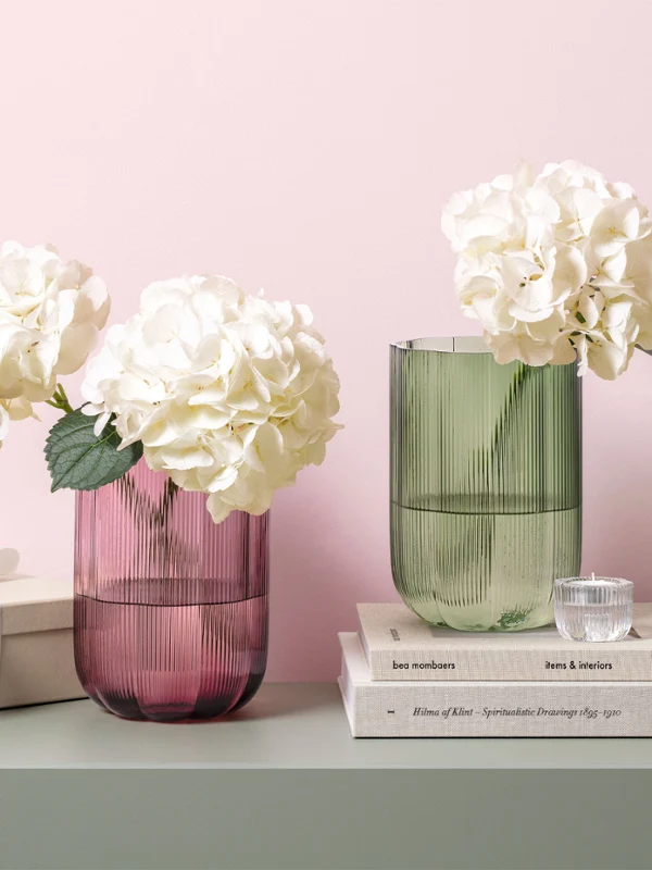 Dos jarrones de cristal Fleur con hortensias blancas sobre una mesa. Dos jarrones de cristal Fleur con hortensias blancas sobre una mesa junto a una pila de libros, una caja de regalo y una pequeña lámpara verde.