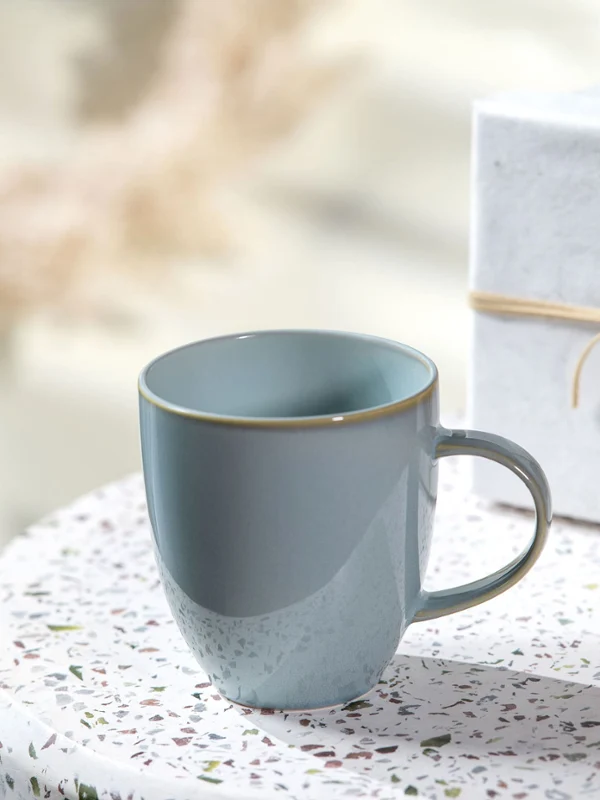 Eine Crafted Tasse auf einem runden Tisch neben einer weißen Geschenkbox mit beiger Schleife und einer runden Karte.