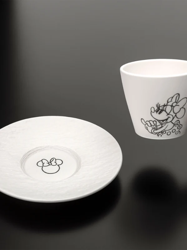 Hand hält eine weiße Manufacture Mickey Tasse mit Untertasse Eine Hand hält eine weiße Manufacture Mickey Tasse mit einer schwarzen Illustration neben einer passenden Untertasse auf einer schwarzen Fläche.