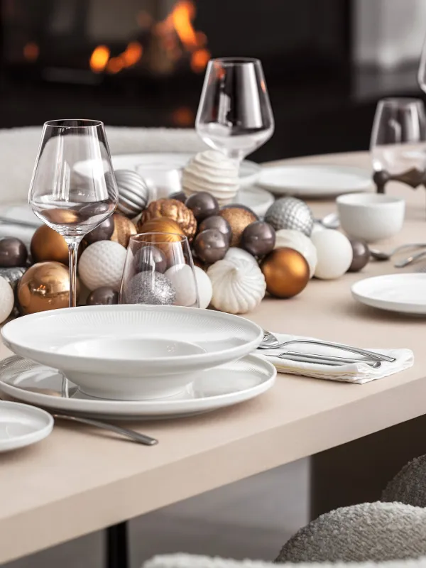 A dining table set with white Afina plates, glasses, candles, and golden Christmas decorations, with a fireplace in the background.