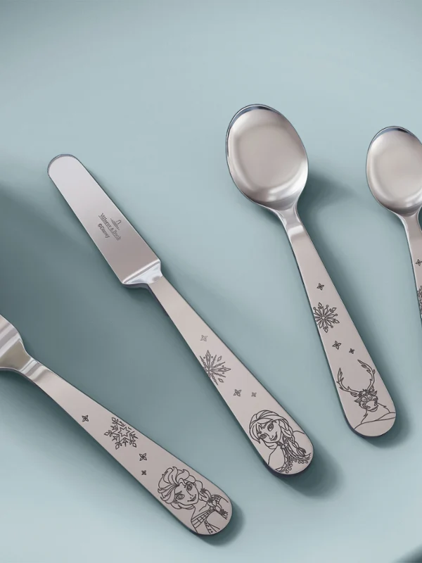 Cubiertos con mangos decorativos de la colección Disney Frozen sobre una mesa azul claro. Un tenedor, un cuchillo, una cuchara y una cucharilla con mangos decorativos de la colección Disney Frozen están dispuestos sobre una mesa azul claro junto a una taza.