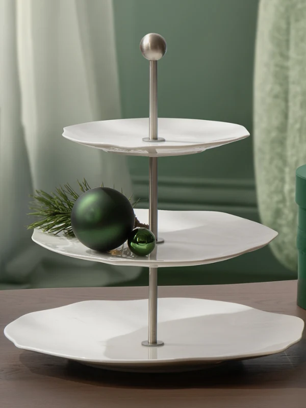 Una bandeja para servir de tres pisos blanca de Pura con un adorno verde y follaje, con dos cajas de regalo verdes redondas al lado sobre una mesa de madera.