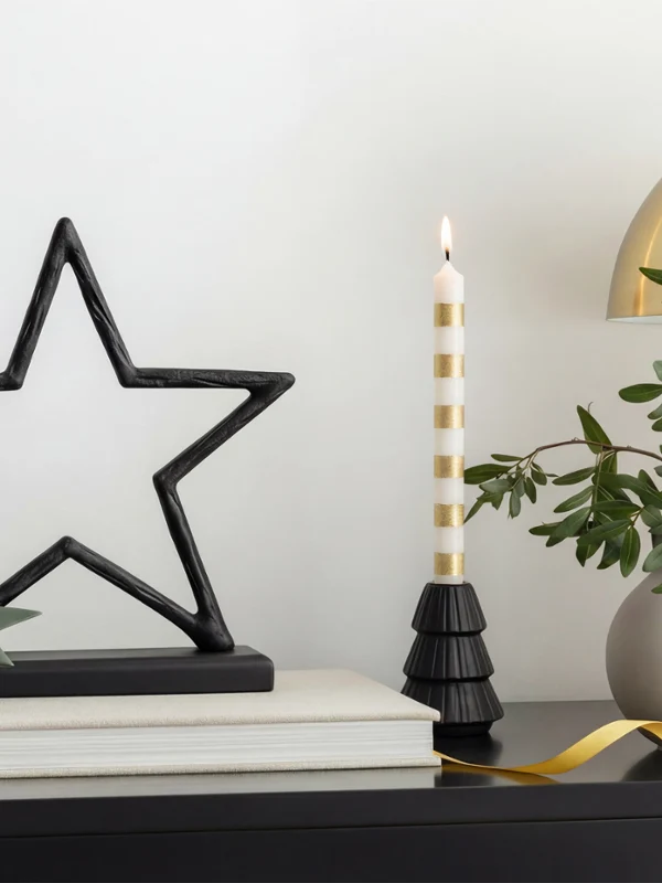 Sobre una mesa con objetos decorativos hay una escultura de una estrella negra, una vela a rayas, una plantita y una lámpara dorada.