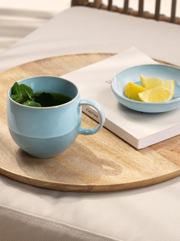 Eine blaue Perlemor Aqua Tasse mit Tee und eine blaue Perlemor AquaSchale mit Zitronenscheiben auf einem Tablett, daneben ein Buch und eine Tüte mit Zitronen.
