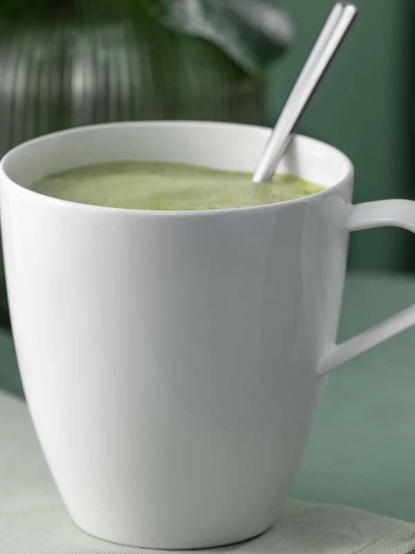 Ein weißer Pura Henkelbecher, gefüllt mit einem grünen Getränk Ein weißer Pura Henkelbecher, gefüllt mit einem grünen Getränk und einem Löffel, der auf einer hellen Fläche steht.
